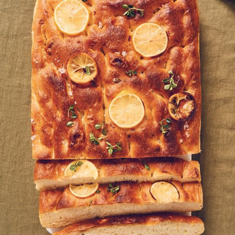 A loaf of bread with lemon slices and herbs baked on top on a mustard-brown tablecloth