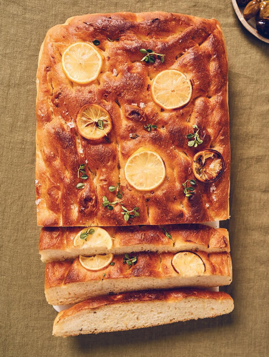 A loaf of bread with lemon slices and herbs baked on top on a mustard-brown tablecloth