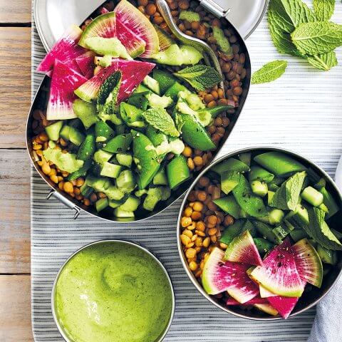A silver bento box and bowl filled with lentils and greens with a bowl of green dressing on the side