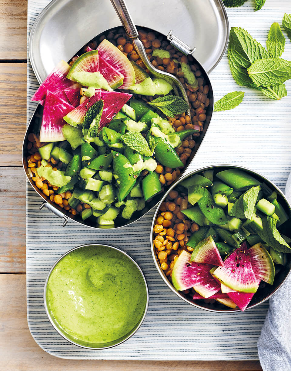 A silver bento box and bowl filled with lentils and greens with a bowl of green dressing on the side