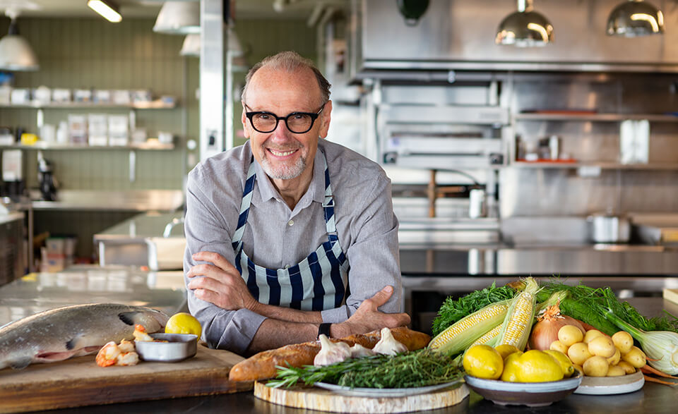 A man, Michael Bonacini, leans on a counter laid with ingredients and smiles