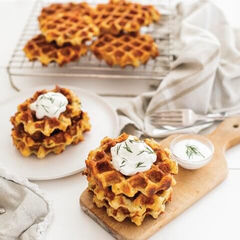 A wooden board with waffles, white sauce and a tray with more visible in the background
