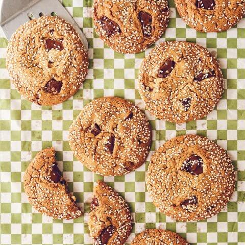 A chocolate chip cookie sprinkled with sesame seeds being lifted with a spatula off a green-and-white checkered sheet of them
