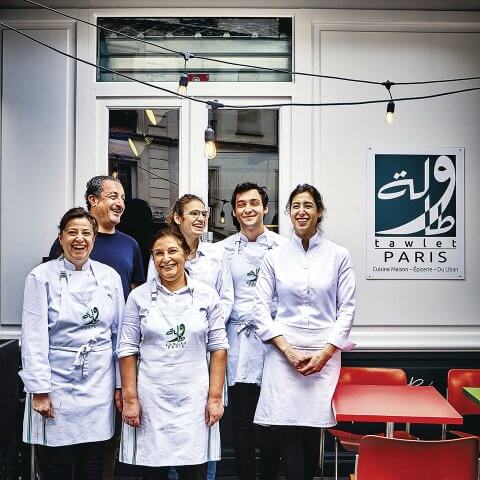 A group of white-clad chefs smiling together in front of a storefront