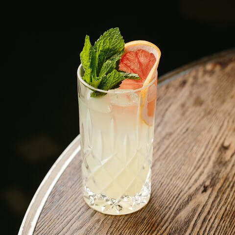 A cocktail garnished with a grapefruit wheel and mint on a wooden table against a dark background