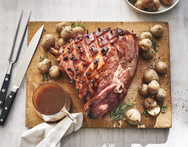Glazed ham on a cutting board with baby potatoes and a small jug of gravy.