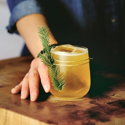 A woman holding a cocktail with an evergreen garnish