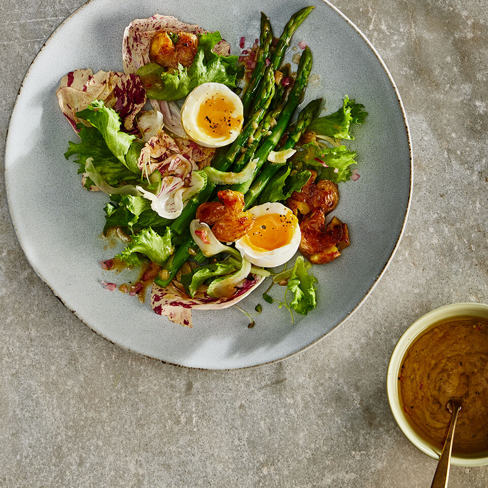 Fresh mixed greens, asparagus, soft-boiled egg and dressing in a small bowl on a grey plate