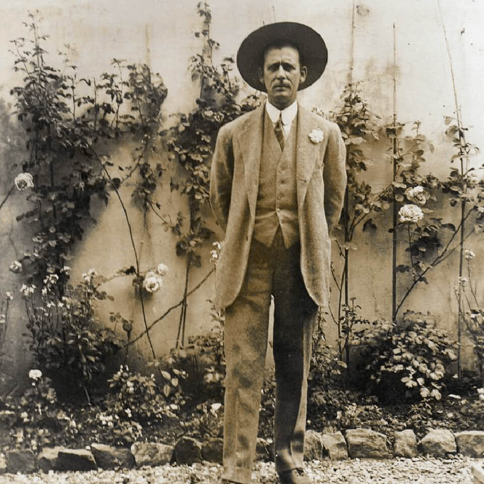 An old sepia-toned photo of a man in a suit and brimmed hat standing in front of a fence with plants