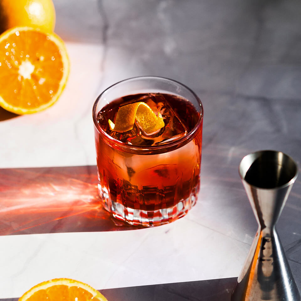 A negroni cocktail on a marble surface with a jogger and oranges next to it