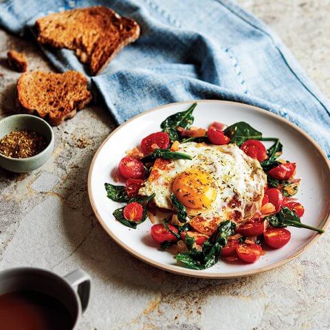 A breakfast dish with fried halloumi and an egg, wilted spinach, tomatoes and halloumi with toast, tea, and a light blue napkin visible in the background