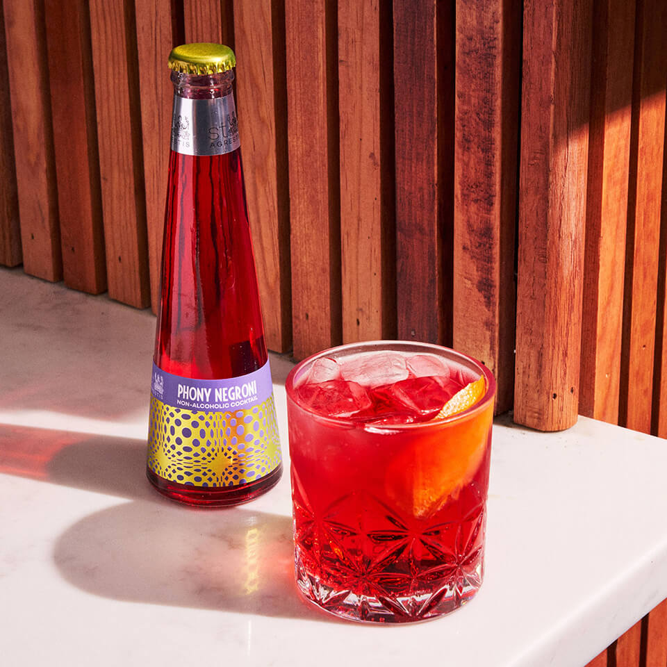 A Negroni in a glass and a bottled one on a white surface with a wooden panelled wall in the background