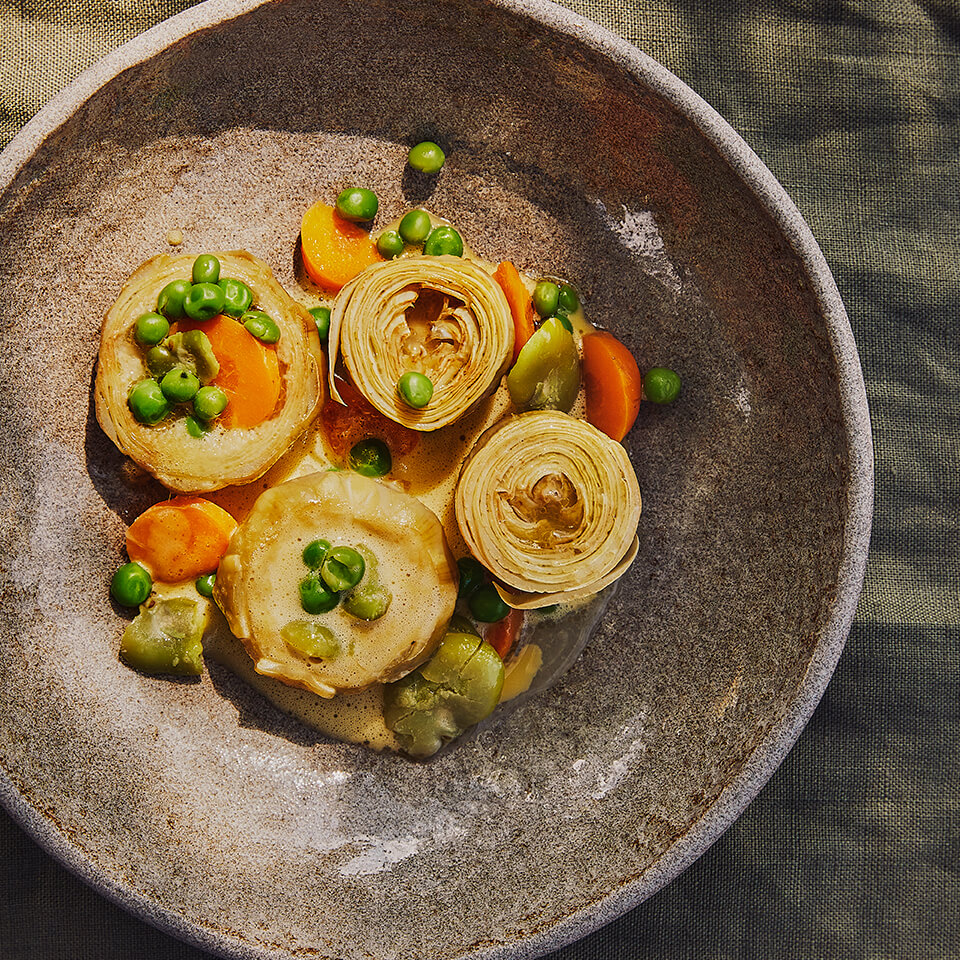 A grey dish with artichokes, peas, other vegetables and sauce