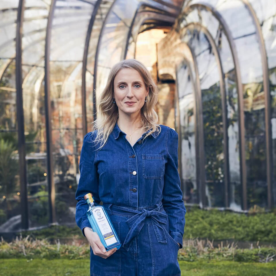 A woman, Dr. Anne Brock, in a denim jumpsuit holding a bottle of Bombay Sapphire in front of a glass atrium