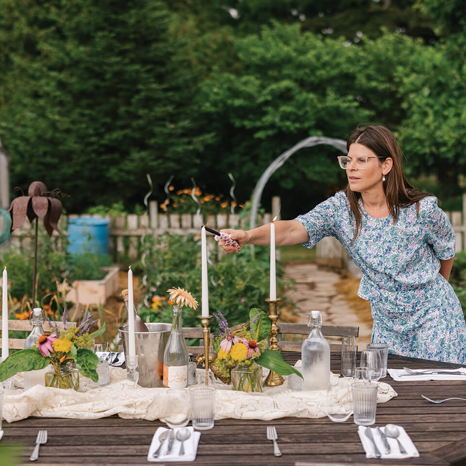 Cookbook author Christine Flynn setting an elegant outdoor dinner table set at golden hour with candles, flowers and seasonal dishes