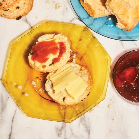 A yellow plate with a currant scone cut in half and spread with butter and jam