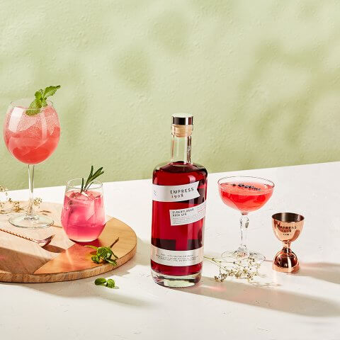 A bottle of pink gin and glasses with various cocktails and a jigger on a white surface with a light green wall visible in the background