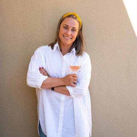 A woman, Geena Luckett, leans against a wall and smiles while holding a glass of wine