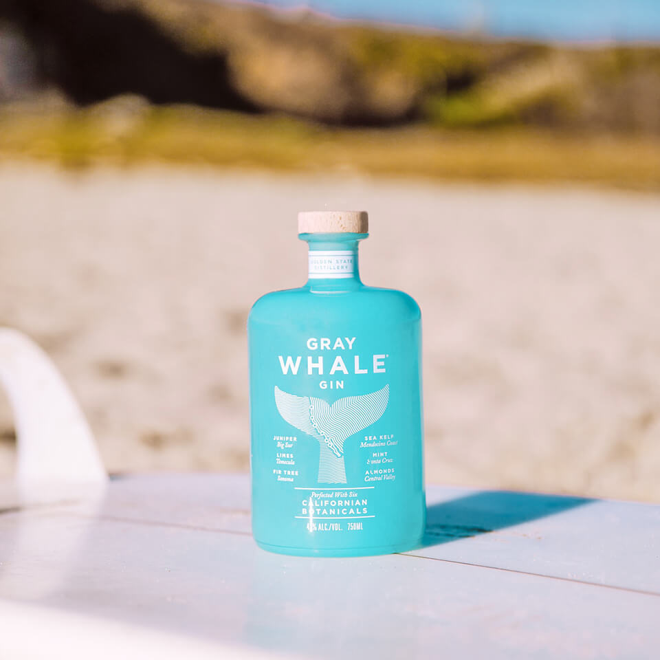 A blue bottle of gin resting on a surfboard with a beach in the background