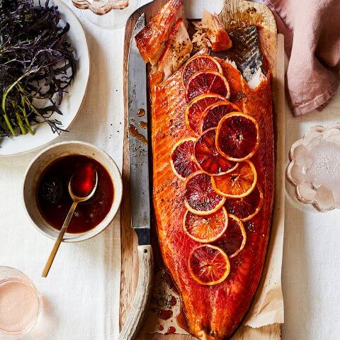 A long board with salmon topped with citrus wheels and a bowl of dressing next to it
