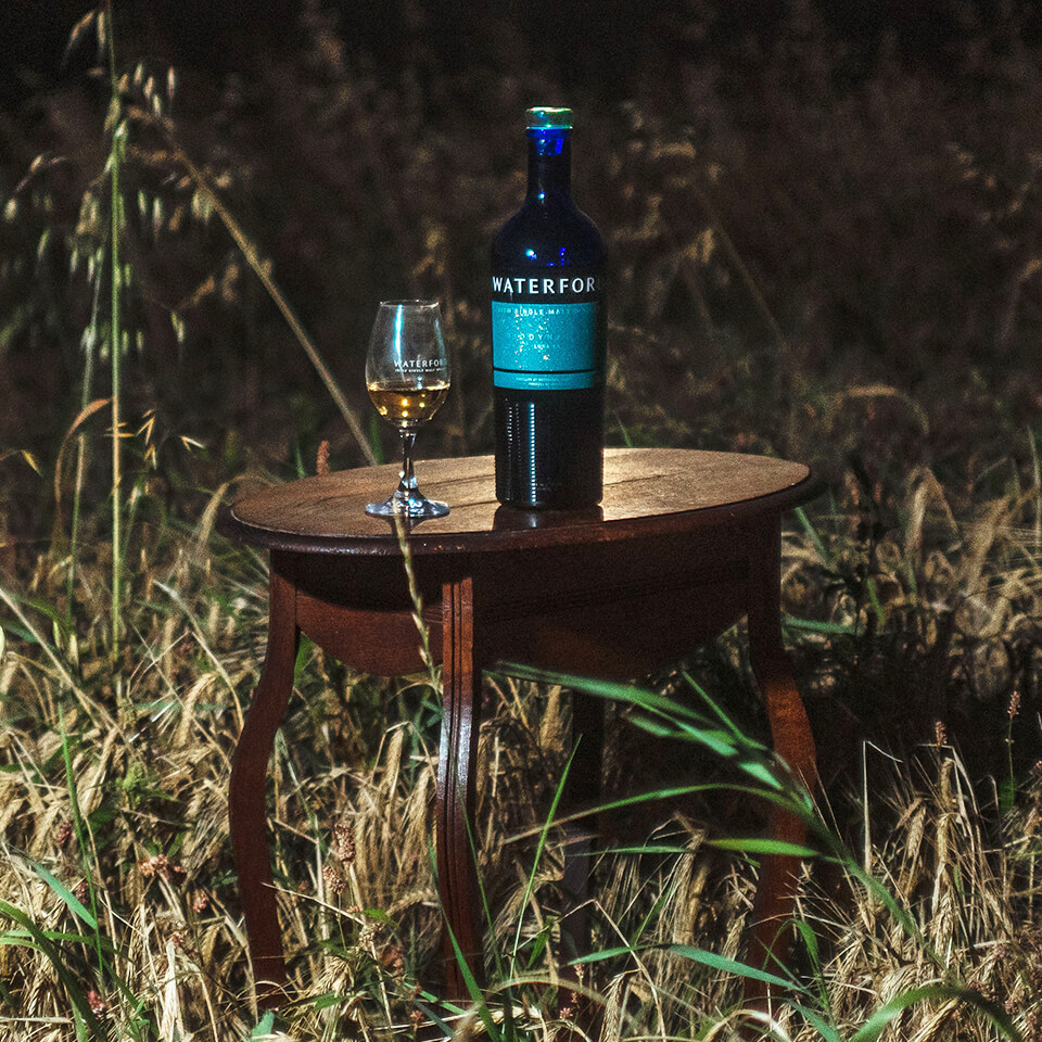 A blue bottle of whisky and a glass on a small wooden table in a field at night