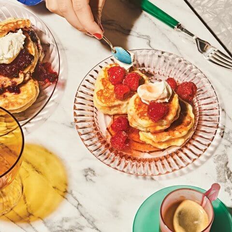 A person's hand holding a spoon over a dish with pancakes and fruit