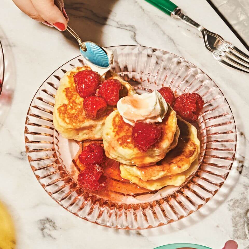 A person's hand holding a spoon over a dish with pancakes and fruit