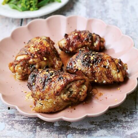 A pink flower-shaped dish with roasted chicken and a white plate with greens visible behind it on a grey surface
