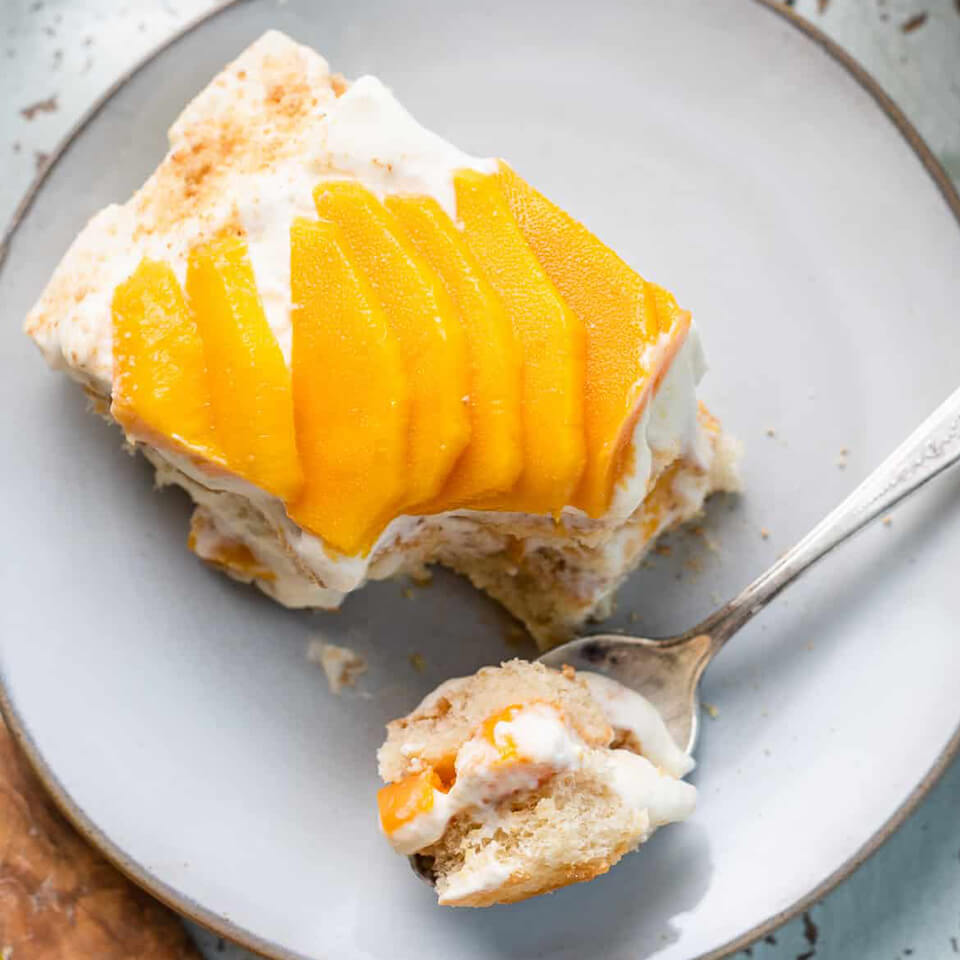 A slice of mango float with a spoon on a plate is an easy mango float recipe has three layers of whipped cream with small mango chunks on top.