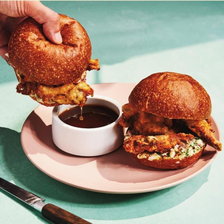 Crispy mushroom French dip sandwiches and a person's hand dipping one on a light pink plate on a light blue surface