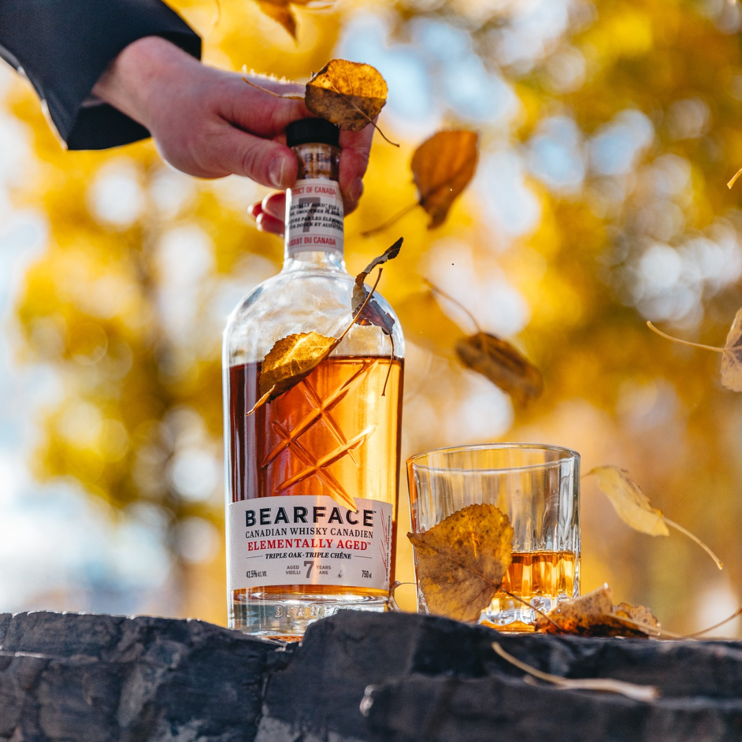 A hand placing a bottle of whisky down on a log beside a glass of whisky.