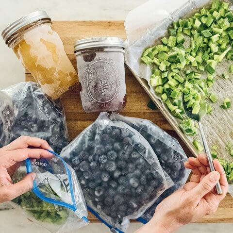 An assortment of fruits and vegetables prepped for freezing on a wooden cutting board, with a person's hands spooning
