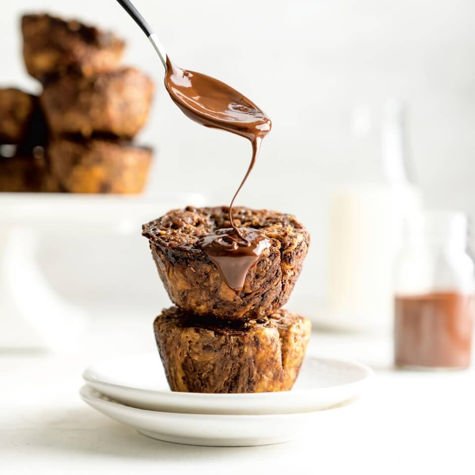 Two chocolate butter tarts on a plate being drizzled with chocolate from a spoon and more butter tarts visible in the background