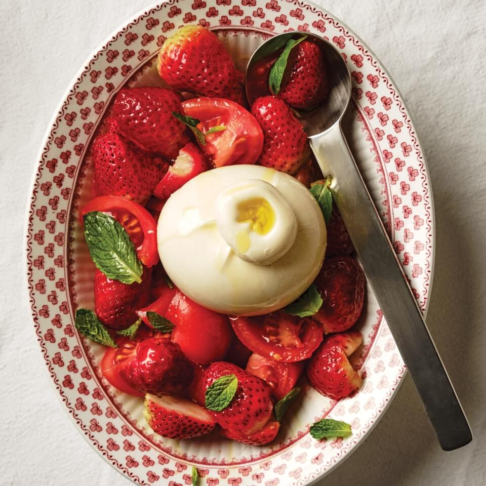 A dish with tomato and strawberry salad topped with burrata, and a serving spoon