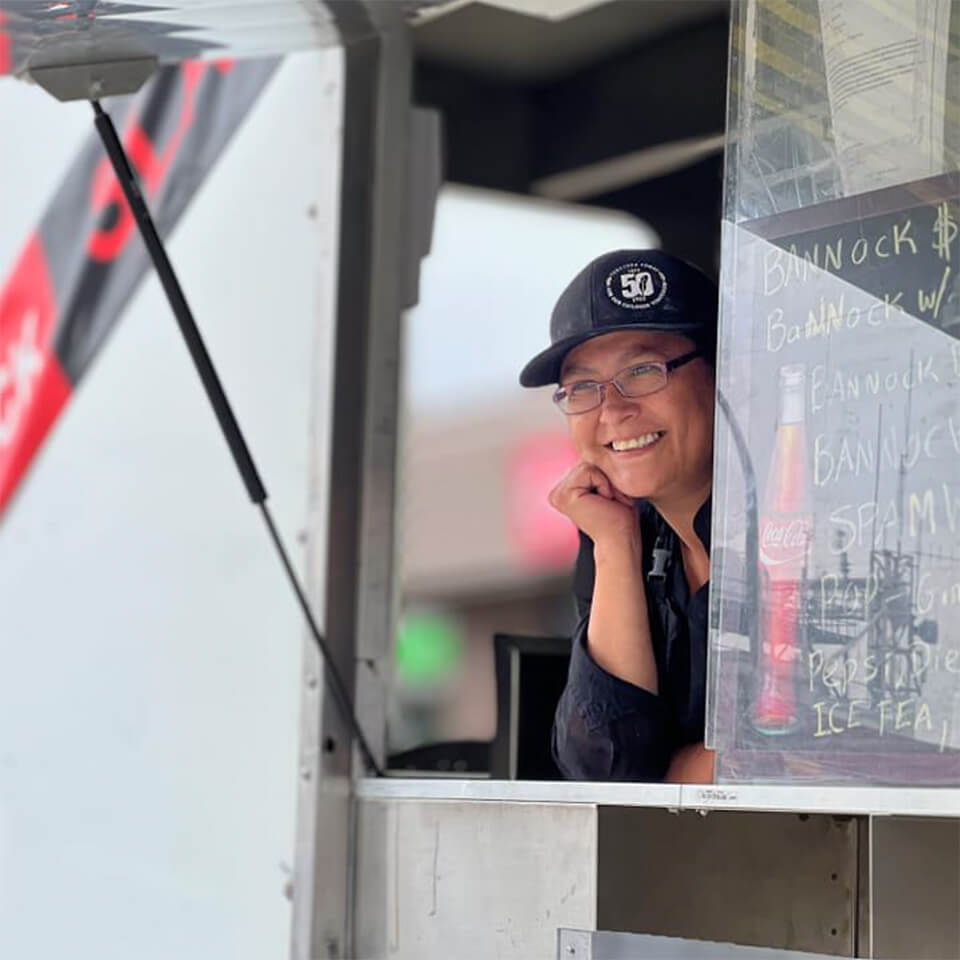 A person smiling in the window of a white food truck with a chalkboard menu listing different bannock items