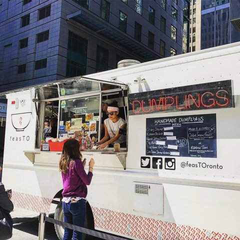 A white food truck with a neon sign reading "DUMPLINGS" and a woman handing food to a customer out the window