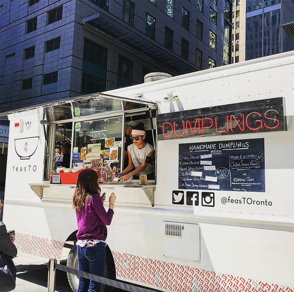 A white food truck with a neon sign reading "DUMPLINGS" and a woman handing food to a customer out the window