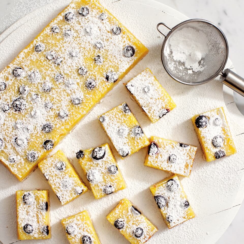 Lemon bars with blueberries dusted with icing sugar and a small sifter