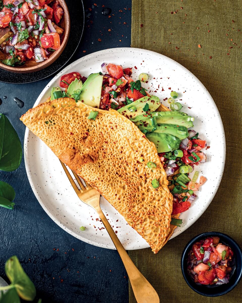 A folded chickpea pancake with a tomato and avocado filling on a white plate with a gold fork. Small bowls of the filling are visible around it