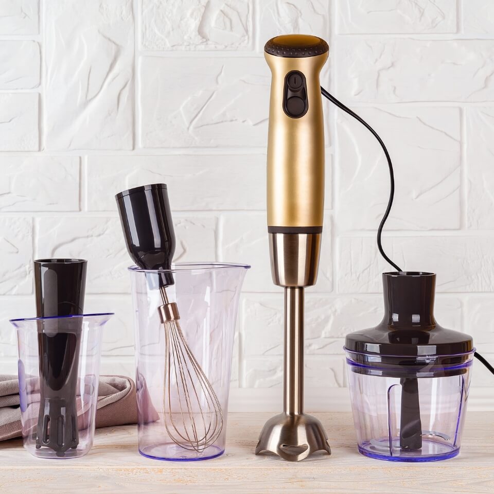 A golden immersion blender with attachments next to it on a countertop