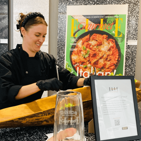 A woman mixing Caesar salad in a large wooden bowl with a cutout of a magazine cover visible behind her