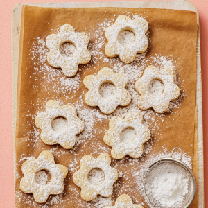 A cookie sheet with flower-shaped Canestrelli Italian Christmas Cookies cookies and dish of powdered sugar on a pink surface