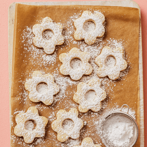 A cookie sheet with flower-shaped Canestrelli Italian Christmas Cookies cookies and dish of powdered sugar on a pink surface