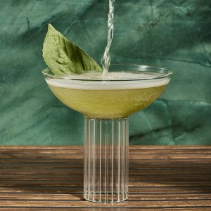 A light green Basil Spritz cocktail being poured into a glass with a basil leaf garnish -