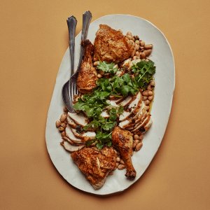 An oval-shaped platter with chicken and beans and cutlery on an orange surface