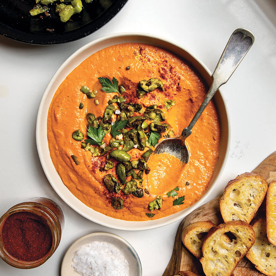 Aerial shot of a bowl of orange dip topped with olives, next to toasted baguette slices, a small bowl of salt and paprika.