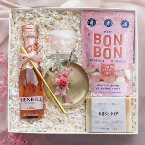 A box with a pink package of candy, bar of soap, candle, bottle of rose and a brass dish on shredded paper.
