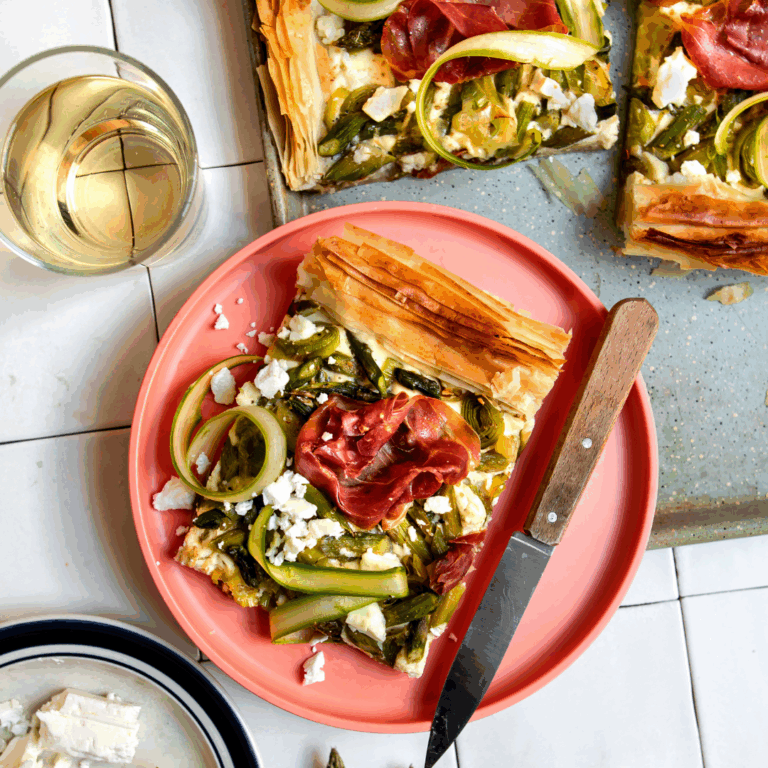 Slice of flaky phyllo tart topped with zucchini ribbons, asparagus, feta and prosciutto on a pink plate, with a knife and glass of white wine nearby.