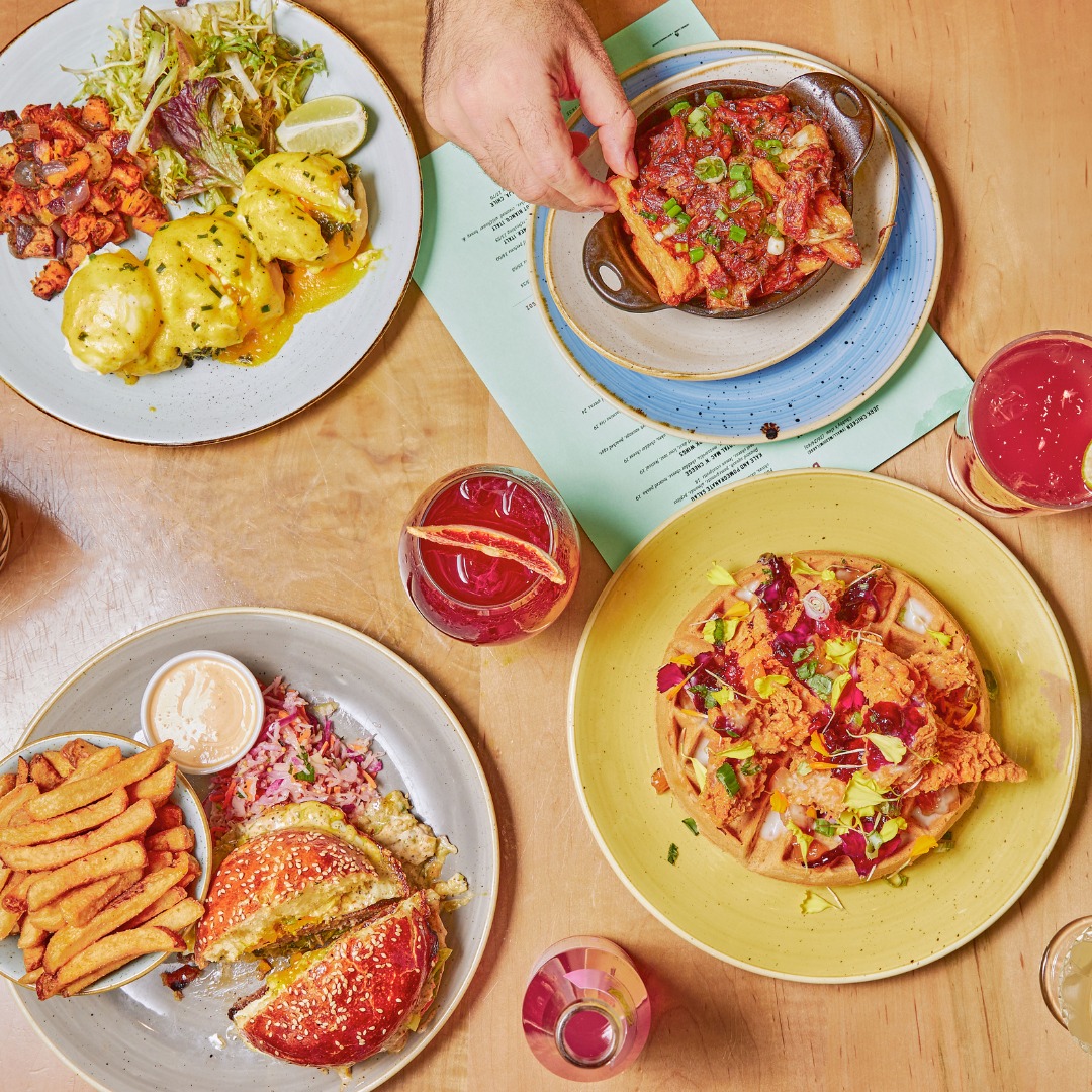 Four plates of brunch dishes on a wooden table with three pink-coloured drinks. 