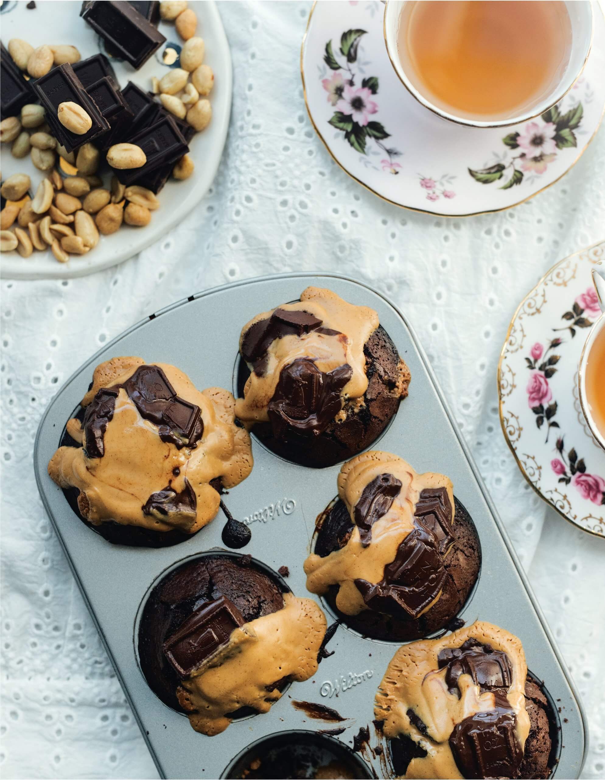 A pan of chocolate muffins on a table with two cups of tea and a plate with peanuts and chocolate.